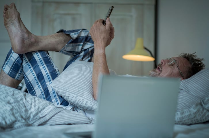 Unhealthy senior man in his 60s with greying beard sleepless in bed while night using laptop and smart phone.